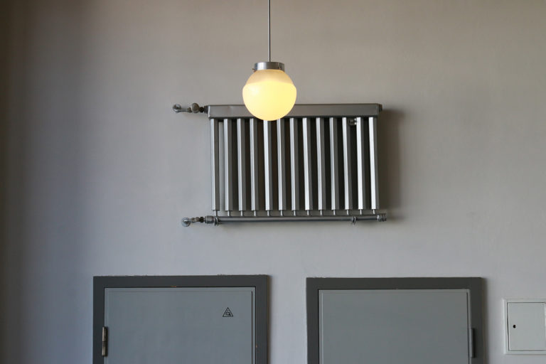 Von der Decke hängt eine runde, leuchtende Lampe herab. Dahinter sieht man einen grauen Heizkörper an der Wand. Darunter zwei angeschnittende, graue Türen