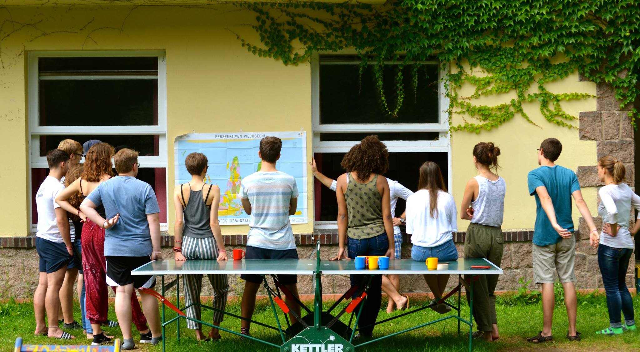 Jugendliche von hinten mit Blick zu einer Hauswand, an der eine umgedrehte Weltkarte hängt mit dem Titel "Perspektiven wechseln"