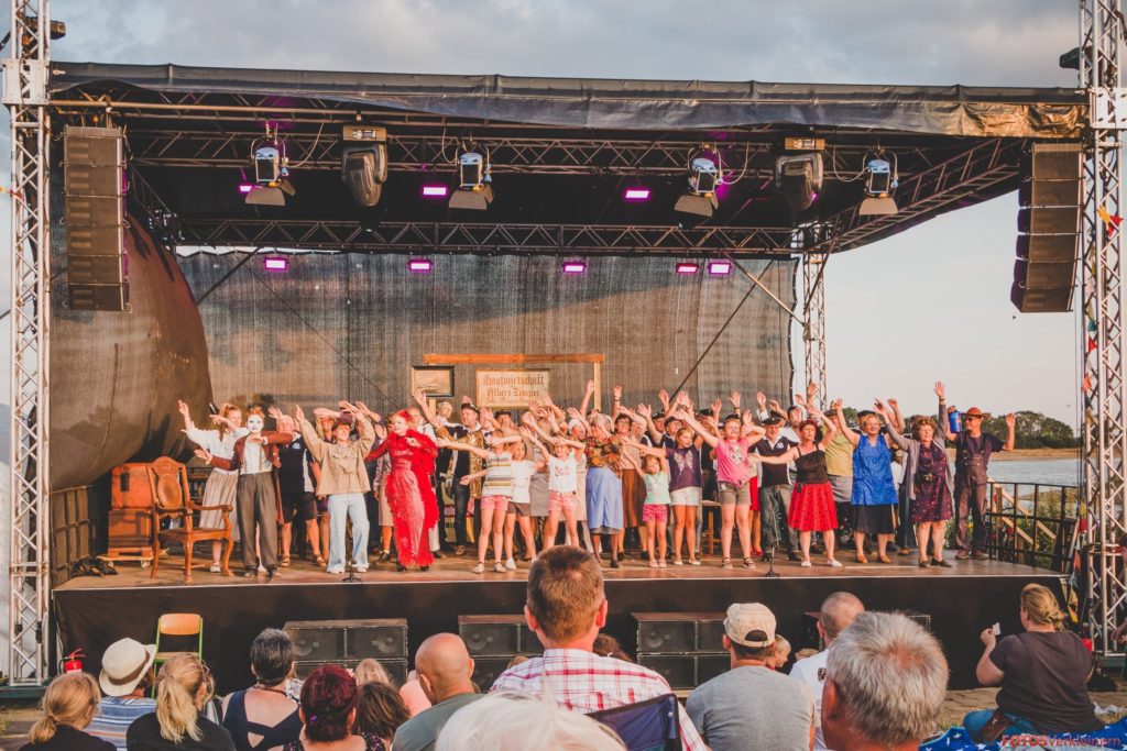 Das Foto zeigt alle Mitwirkenden des Theaterstücks auf der Bühne. Die Gruppe schwingt die Arme über den Köpfen.