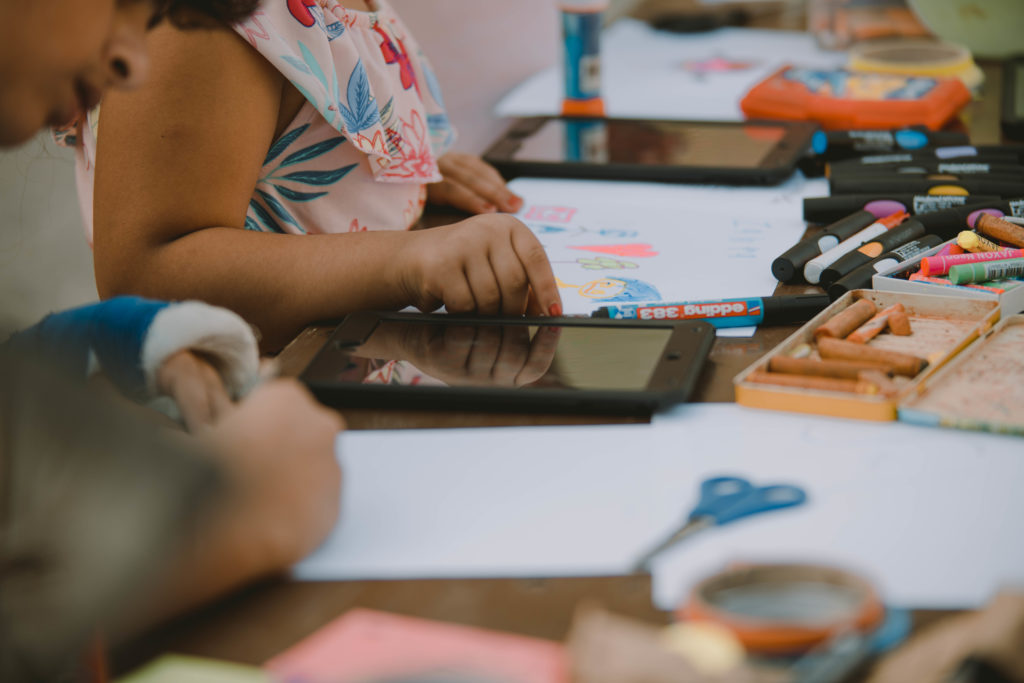 Man sieht die Hände von Kindern beim Malen. Vor ihnen liegen zahlreiche Stifte, Wachsmaler und Papier. Außerdem liegen zwei Tablets auf dem Tisch.