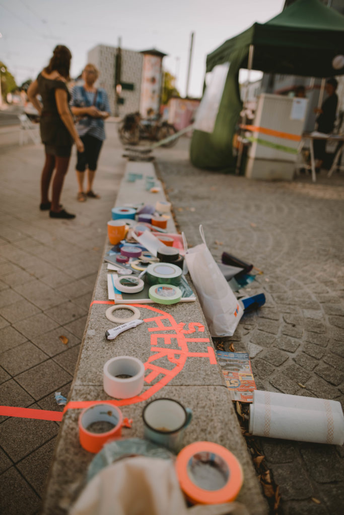 Auf einer flachen Steinmauer liegen viele verschiedene Rollen von buntem Klebeband. Mit orangenem Klebeband ist ein Sitzplatz abgeklebt und darauf steht "Sit here". Im Hintergrund stehen 2 Personen, die sich unterhalten und ein grüner Pavillon.