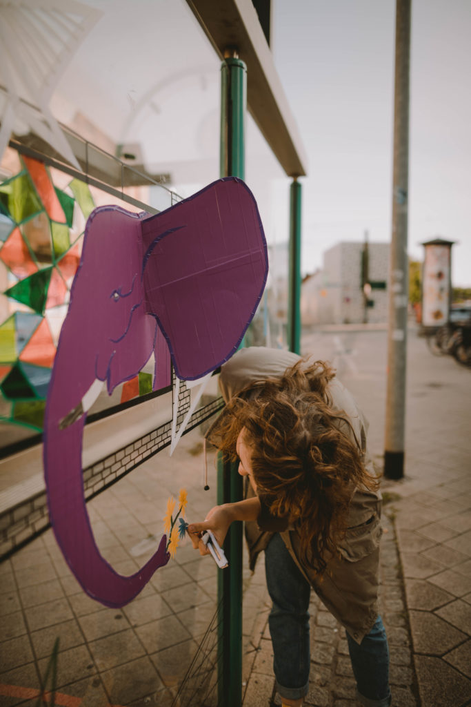 Eine junge Frau beklebt die Scheibe einer Straßenbahnhaltestelle mit verschieden farbigem Klebeband. Auf der Scheibe entsteht ein 3D-Elefantenkopf. Das Bild entstand im Rahmen der Straßengalerie in Magdeburg Südost.