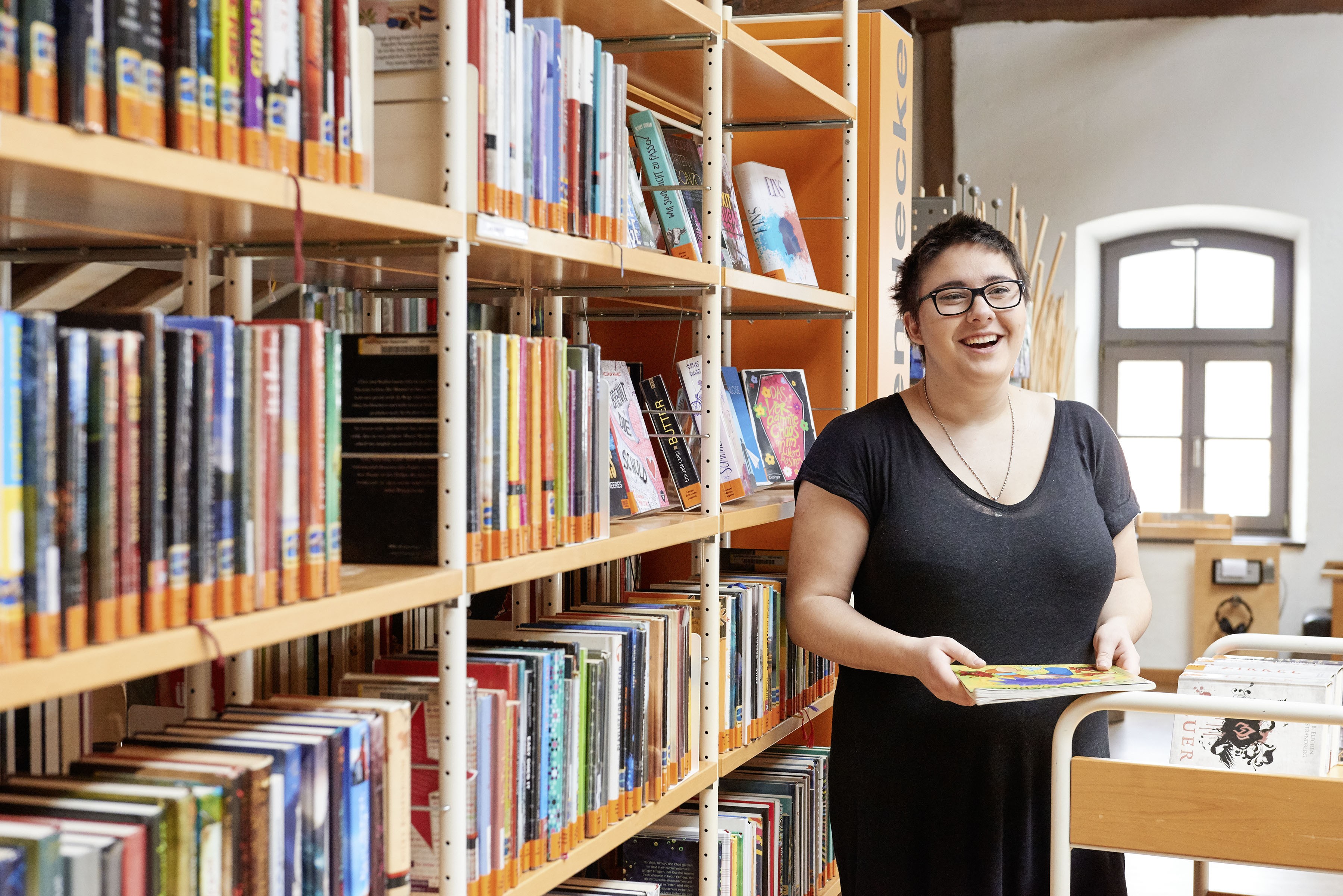 Eine junge Frau steht vor einem Bücherregel in der Bibliothek. Vor ihr steht ein Bücherwagen und sie hält ein Buch in der Hand, was sie einsortieren will. Sie lächelt an der Kamera vorbei jemanden an.