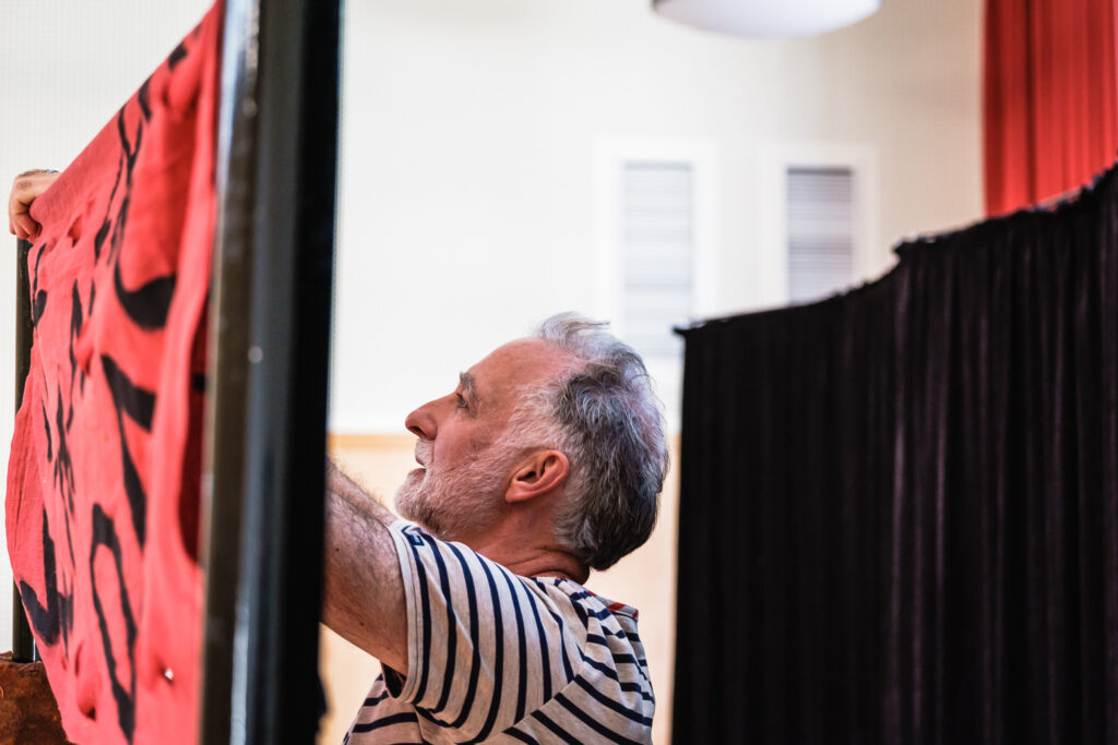 Ein älterer Mann steht an einer Leinwand und spannt einen Stoff auf. Hinter ihm sieht man einen dunklen Theatervorhang.