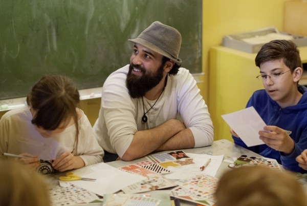 Ein junger Mann sitzt an einem Tisch voller Papier und Materialien. Neben ihm sitzen Kinder, die etwas basteln und malen. Er schaut nach links aus dem Bild raus und lächelt.