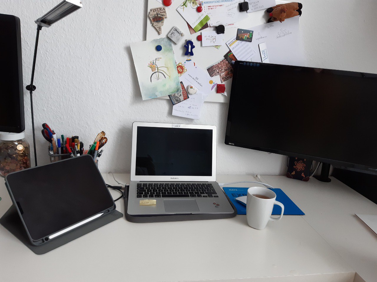 Bild eines Schreibtischs mit mehreren Bildschirmen und einem Laptop. Davor steht eine Kaffeetasse. Im Hintergrund an der Wand hängen ein paar Notizen und Bilder an einer Pinnwand.