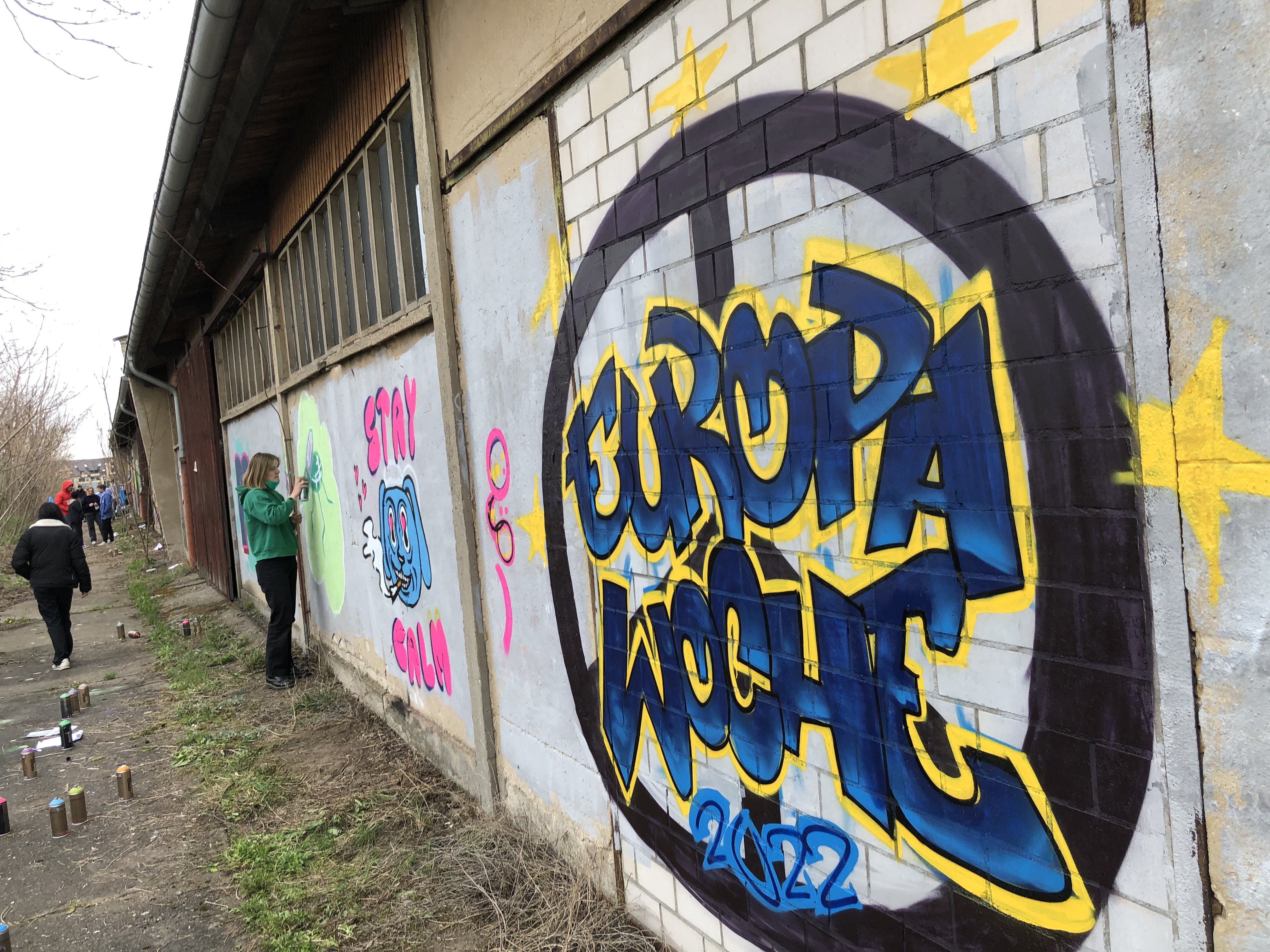 "Europawoche" steht in Blau und Gelb gesprüht an einer Wand. Im Hintergrund sind mehr Personen zu sehen, die ebenfalls etwas an die Wand sprühen.