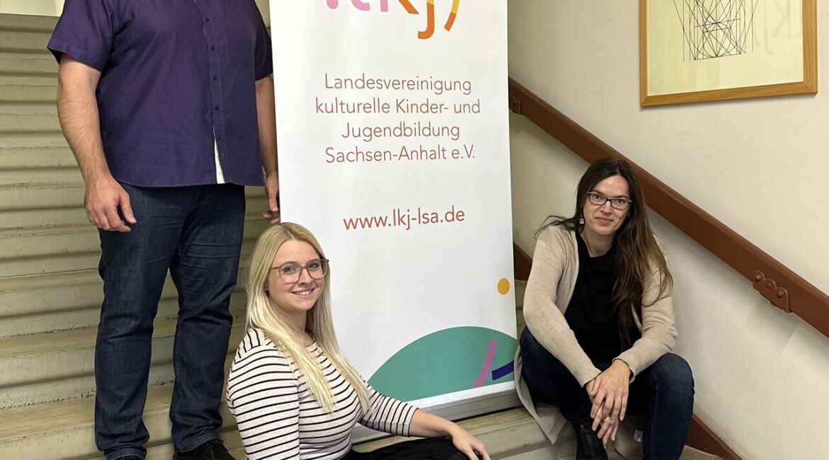Drei Mitglieder des neuen Vorstands der LKJ Sachsen-Anhalt. Katrin Brademann, John Palatini und Rebecca Hempel sitzen auf einer Treppe im Flur der LKJ Sachsen-Anhalt.