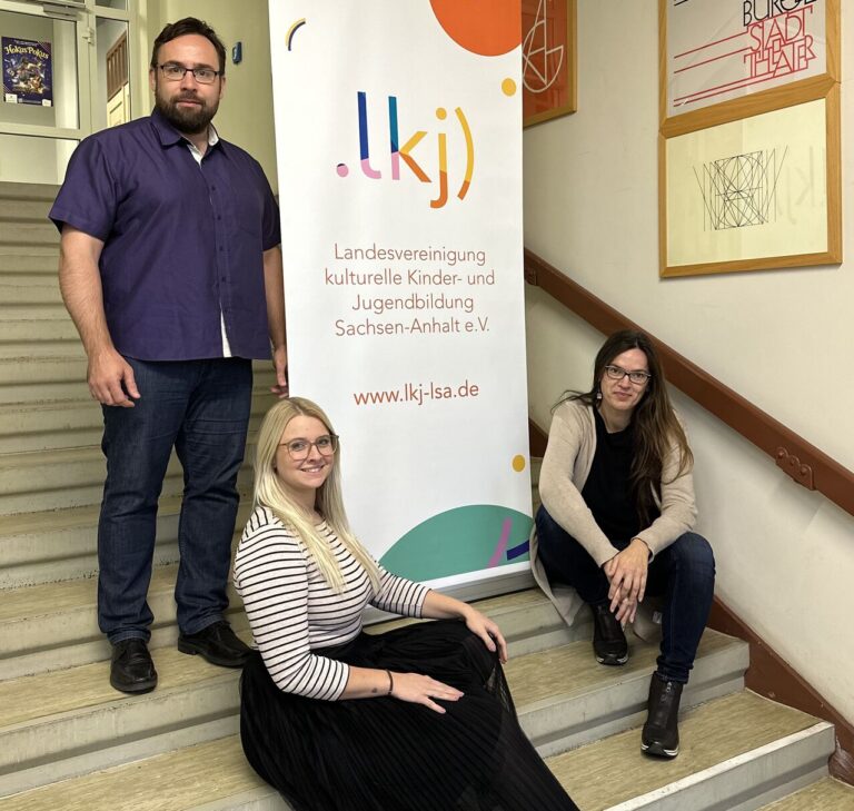 Drei Mitglieder des neuen Vorstands der LKJ Sachsen-Anhalt. Katrin Brademann, John Palatini und Rebecca Hempel sitzen auf einer Treppe im Flur der LKJ Sachsen-Anhalt.